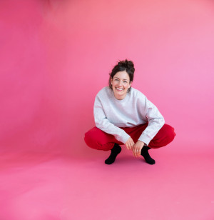 Lena freundlich lächelnd mit grauem Pullover und roter Jogginghose in Hocke auf pinkem Hintergrund.