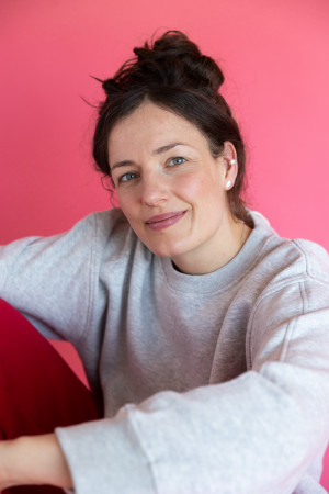 Lena mit grauem Pullover und roter Jogginghose, ihr rechts Knie an sie herangezogen, in die Kamera schauend