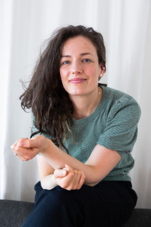Lena mit grünem, strukturreichem T-Shirt. Die Hand zugewandt in die Kamera gerichtet.